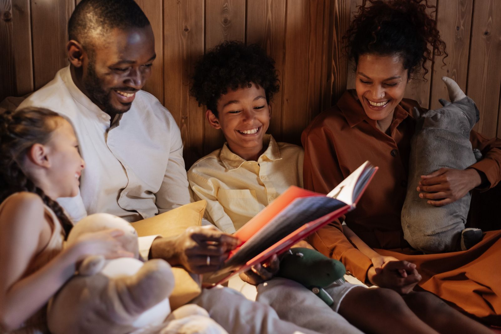 A family of four sits close together smiling as the father reads from an open storybook in warm evening light.