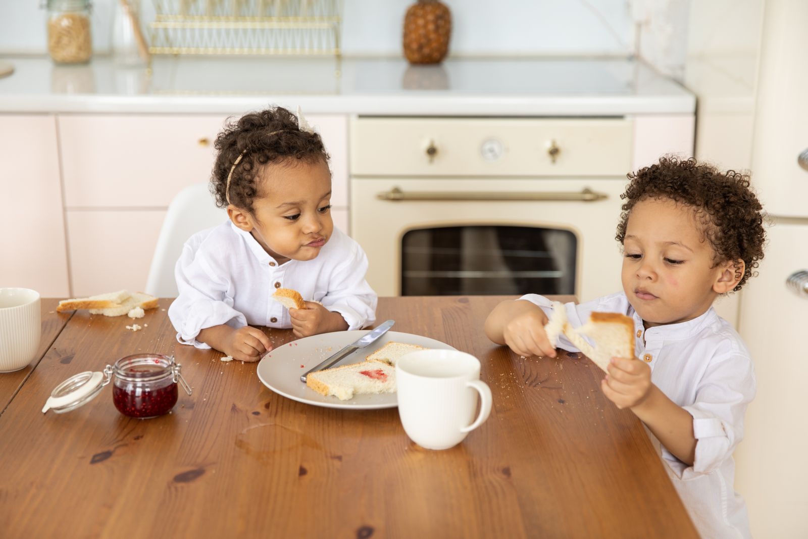 Two young children sharing bread and jam at a wooden kitchen table at home